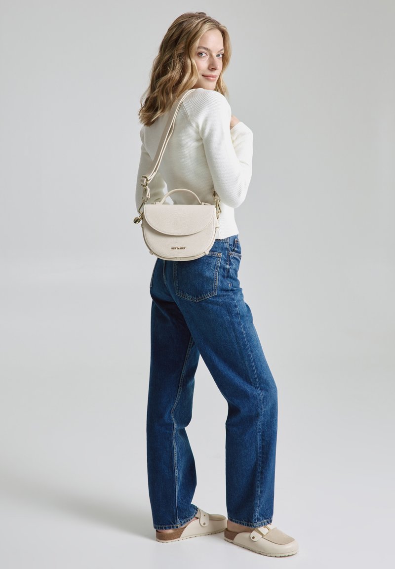 Beige strukturierte Handtasche mit einem runden oberen Teil, verstellbarem Trageriemen und strukturierter Oberfläche. Das Model trägt einen weißen Pullover und blue jeans.