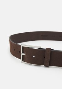 Curea din suede maro cu o cataramă din metal argintiu, având o textură netedă și multiple găuri pentru ajustare. Marca este vizibilă pe interior.
