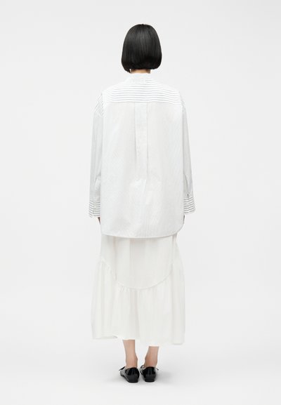 Άτομο με κοντά μαύρα μαλλιά, γυρισμένο προς τα πίσω, που φοράει φαρδιά λευκή ριγέ πουκάμισο, μακριά λευκή φούστα και μαύρα επίπεδα παπούτσια, σε απλό φόντο.