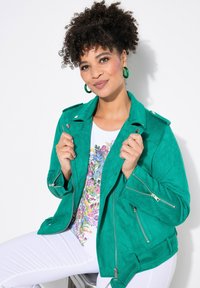 Giacca da moto in suede verde con tasche con zip, abbinata a una maglietta floreale e pantaloni bianchi. Modello in posa seduta.