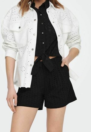 Vrouw draagt een zwart eyelet-knop-gebonden shirt en bijpassende korte broek, gelaagd met een witte eyelet-knopjas met opgerolde mouwen.