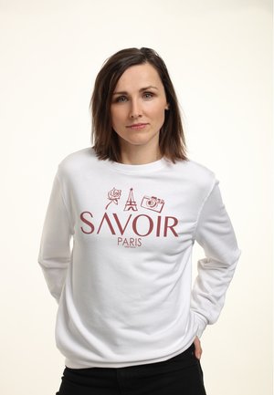 Femme aux cheveux bruns portant un sweat-shirt blanc avec le texte rouge « SAVOIR PARIS » et des icônes de rose, Tour Eiffel et appareil photo.