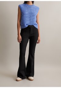 Femme vêtue d’un top en tricot bleu sans manches, d’un pantalon noir évasé taille haute et de baskets blanches, debout devant un fond uni.