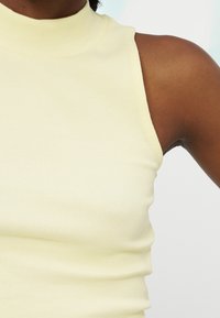 Gros plan sur une personne portant un haut sans manches côtelé jaune pâle à col montant, sur un fond clair.