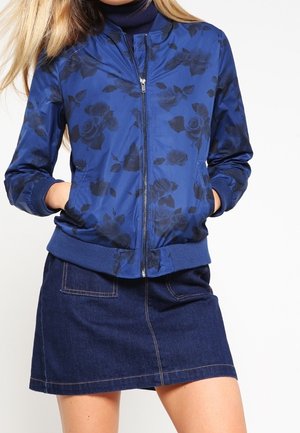 Kobieta w niebieskiej, kwiecistej bomberce zapinanej na zamek oraz ciemnej spódnicy jeansowej z przednimi naszywanymi kieszeniami, z rękami w kieszeniach kurtki.