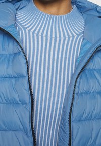 Niebieska pikowana kurtka z zamkiem błyskawicznym, nałożona na jasnoniebiesko-biały sweter w paski z prążkowanym dekoltem.