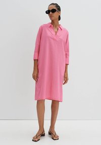 Rosa Leinenkleid mit Kragen, Dreiviertelärmel und seitlichen Schlitzen. Getragen mit schwarzen Sonnenbrillen und Sandalen mit Riemen.