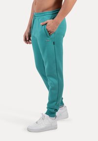 Pantalones de chándal turquesa con cinturilla elástica, bolsillos laterales y una etiqueta con logotipo. Combinados con zapatillas blancas, que presentan un material texturizado.