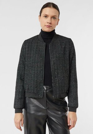 Frau mit dunklem Haar, die eine schwarze, strukturierte Jacke mit Reißverschluss über einem schwarzen Rollkragenpullover und schwarze Lederhosen trägt, vor einem schlichten Hintergrund stehend.