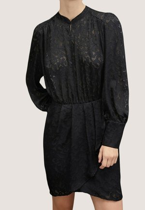 Zwarte jurk met lange mouwen met subtiel bloemenpatroon, ingesnoerde taille, hoge halslijn en omslagmouwen, gedragen door een persoon met hand op de heup.