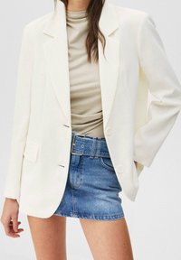 Biały oversize'owy żakiet z wykładanymi klapami, dwoma guzikami i przednimi kieszeniami, zestawiony z beżową bluzką i niebieską dżinsową mini spódnicą.