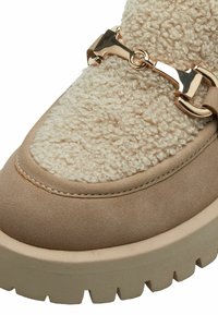 Beige mokkanahka kengät, joissa on teksturoitu kermaisen valkoinen karvapala, kultaketju yksityiskohtana ja paksu, karu pohja. Pyöreä kärki muotoilu.