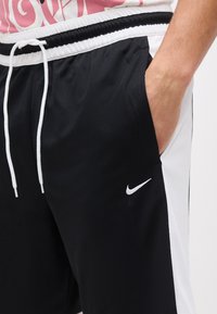 Κοντινό πλάνο ενός ατόμου που φοράει μαύρα και άσπρα σόρτς Nike, με το δεξί του χέρι στην τσέπη και λευκά κορδόνια στη μέση.