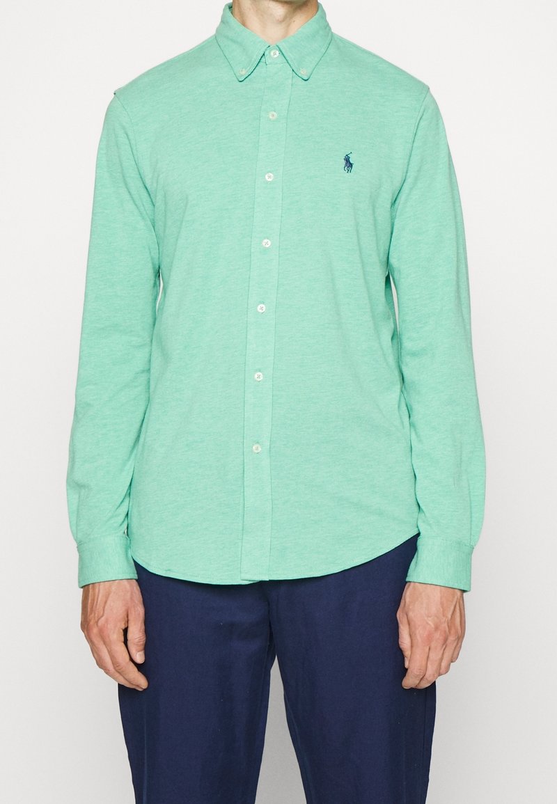 Man wearing mint green long-sleeve button-down shirt with collar and navy blue pants, standing against a plain white background.