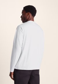 Long-sleeve white top made of smooth fabric, featuring a crew neck and relaxed fit. No visible patterns or accents. Black pants are worn.