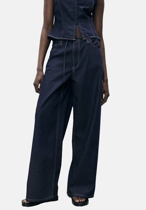 Donkerblauwe denim broek met wijde pijpen, gedragen met een bijpassend mouwloos topje en zwarte sandalen, getoond van taille tot voeten bij een staand persoon.