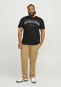 T-shirt en coton noir avec logo de marque blanc, associé à un pantalon cargo beige avec poches latérales et des baskets blanches avec des rayures noires.
