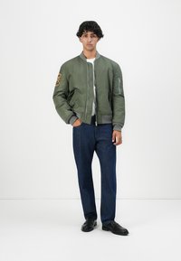 Jonge man die staat en een olijfgroene bomberjack, wit shirt, donkerblauwe jeans en zwarte schoenen draagt, tegen een eenvoudige witte achtergrond.