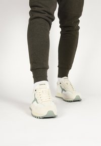Schoenen: witte sneakers met lichtgroene accenten, van suede en mesh materialen, ronde neus, geconfigureerde zool, logo op de tong, gedragen met donkere trainingsbroeken.