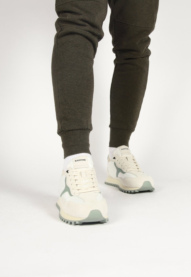 Schoenen: witte sneakers met lichtgroene accenten, van suede en mesh materialen, ronde neus, geconfigureerde zool, logo op de tong, gedragen met donkere trainingsbroeken.