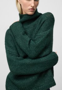 Zielony sweter z dzianiny z wysokim golfem i oversized'owymi rękawami. Tekstura jest miękka i gruba, z prążkowanymi akcentami i luźnym krojem.