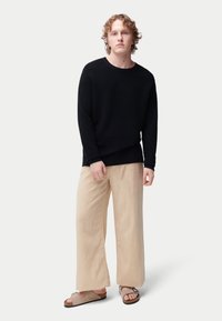 Pull en maille noire texturée, pantalon ample beige clair et sandales marron. Design simple avec une coupe décontractée et une quincaillerie minimale.