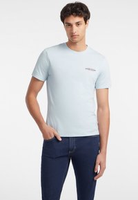 T-shirt bleu deschis cu gât rotund din material bumbac, având un mic logo text în roșu și negru pe partea stângă a pieptului. Mânici scurte, croi lejer.