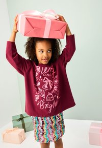 Bordeauxrotes langärmeliges Hemd mit festlichem Grafikdruck, kombiniert mit einem mehrfarbigen Paillettenrock. Hält eine pinke Geschenkbox mit Schleife.