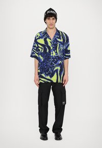 Trumpas rankovėmis marškiniai su mėlynu ir neoniniu žaliu abstrakčiu raštu, derinami su juodais cargo kelnėmis ir juodais batais. Juodas beisbolo kepuraitė su logotipu.