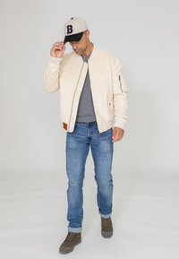 Veste bombardier crème avec des poignets côtelés et une poche zippée, t-shirt gris, jeans bleus et bottes marron. Casquette de baseball en beige et noir.