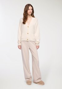 Kremowy sweter z dzianiny z dekoltem w serek, czterema złotymi guzikami i dwiema przednimi kieszonkami. Połączony z luźnymi, jasnobeżowymi spodniami i butami wsuwanymi.