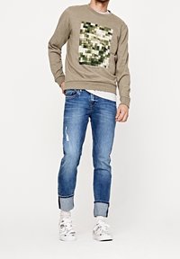Felpa khaki con grafica verde pixelata, abbinata a jeans chiari strappati e sneakers bianche. Vestibilità rilassata e design con collo a giro.