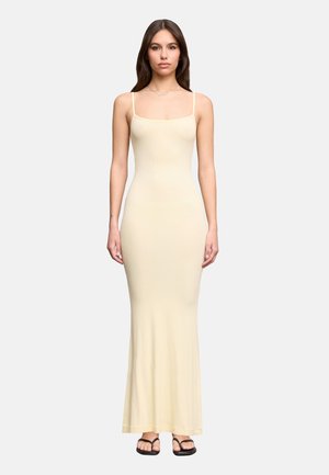 Donna con un abito lungo aderente senza maniche color crema con sottili bretelle e infradito nere, in piedi su uno sfondo neutro.