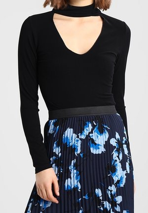 Kvinde iført en sort bluse med lange ærmer og dyb V-udskæring samt en blå, blomsterprintet plisseret nederdel.