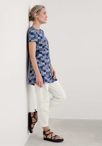 Mėlynos ir baltos raštuotos tunikos viršus su trumpomis rankovėmis, derinamas su baltais kelniais ir juodomis diržinėmis šlepetėmis, atsirėmęs į sieną.