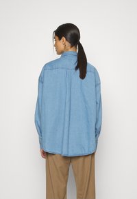 Camicia di jeans azzurro chiaro con vestibilità rilassata, dotata di colletto a punta, maniche lunghe e dettagli di cucitura, indossata sopra pantaloni marroni.