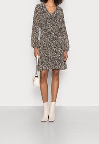 Robe noire à motif floral blanc, col en V, manches longues, taille cintrée. Associée à des bottines blanches et un petit sac à main blanc.