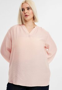 Vaaleanpunainen pitkähiha blouse, jossa on V-pääntie, valmistettu tekstuurista kankaasta. Siinä on rento leikkaus ja hienovaraisia ompelutietoja.