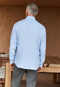 Lichtblauw overhem met lange mouwen in een gestructureerde stof, gecombineerd met grijze broek. Het overhemd heeft knoopmanchetten en een losse pasvorm, getoond vanaf de achterkant.