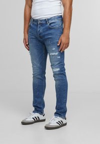 Mees, kellel on seljas kitsas muudetud sinine teksapaar, millel on värvipritsmed, ja valged Adidas tossud mustade triipudega.