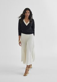 Black v-neck sweater with white trim, paired with a pleated cream maxi skirt and gold heeled sandals, featuring a textural sheen.