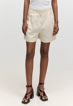 Crèmekleurige geplooide shorts van lichte stof, met een brede tailleband en een knielange snit, gedragen met donkere sandalen met hakken en bandjes.