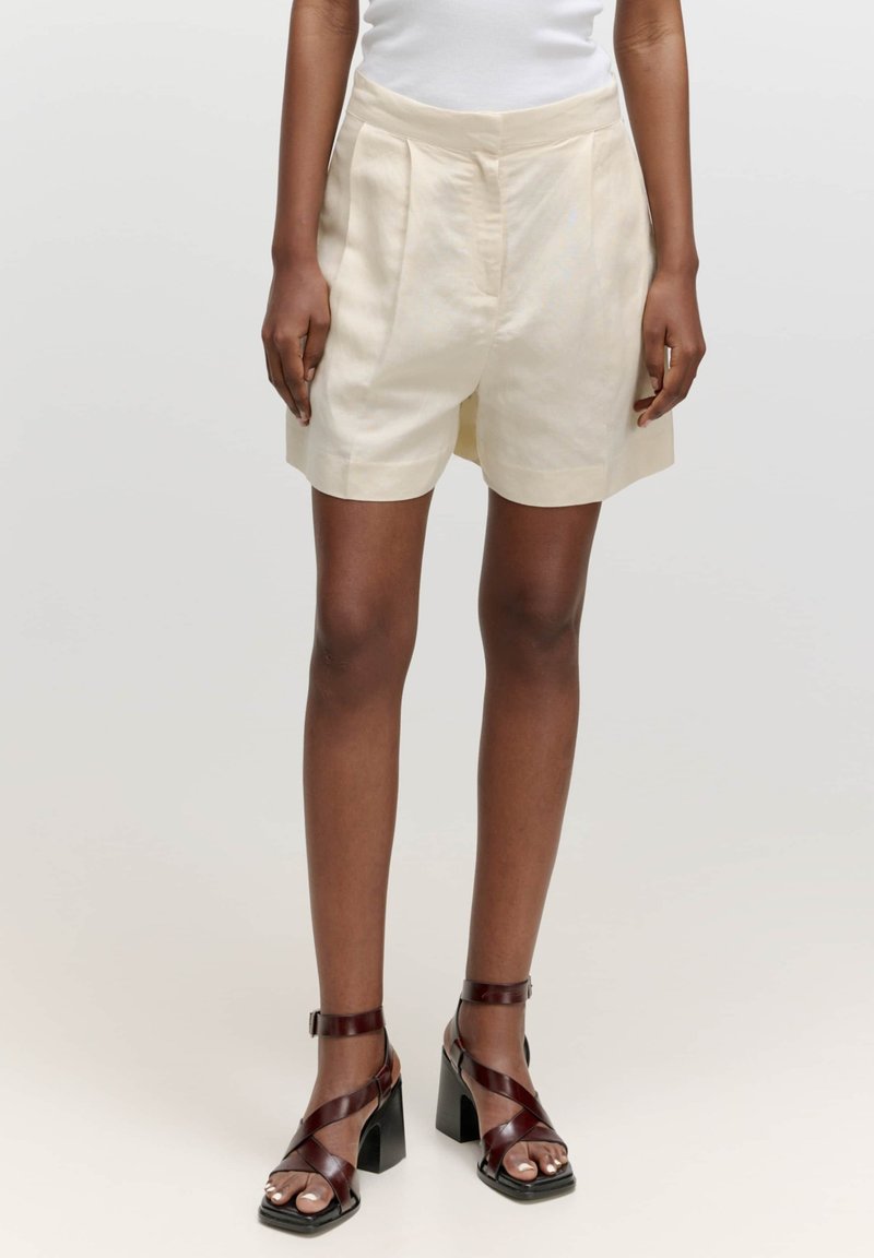 Cremefarbene, plissierte Shorts aus leichtem Stoff, mit einem breiten Bund und knielangem Schnitt, kombiniert mit dunklen, geschnürten Sandalen mit Absatz.