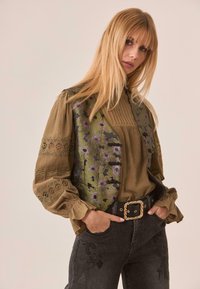 Blusa verde oliva con chaleco bordado floral, mangas abullonadas y tejido texturizado; combinada con jeans oscuros con costura floral.