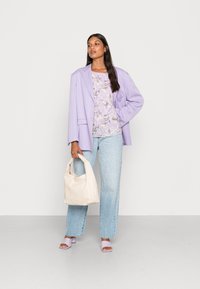 Blazer oversized em lilás sobre uma blusa com padrão floral, jeans de cintura alta em azul claro e sandálias de bloco em lavanda, segurando uma bolsa creme.
