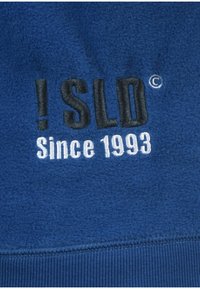 Blå fleece-stof med broderet tekst "! SLD" i sort og "Since 1993" i hvid; har en ribbet kant forneden.