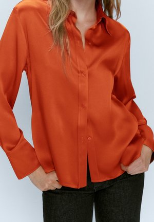 Femme portant une chemise en satin orange vif à boutons avec manches longues, associée à un pantalon sombre, les mains dans les poches.
