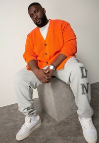 Orangene gerippte Strickjacke mit Knöpfen, graue Trainingshose mit dem Aufdruck "NDN" in fetter Schrift und weiße Sneakers. Modell sitzt auf einem Betonblock.