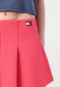 Jupe plissée rose en tissu texturé avec un patch en cuir de la marque Tommy Jeans. Le design présente une taille ajustée et un ourlet évasé.