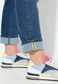 Jeans de mezclilla azul medio con dobladillos acampanados, con un acento en verde neón. Combinados con zapatillas multicolor en tonos pastel y blanco.